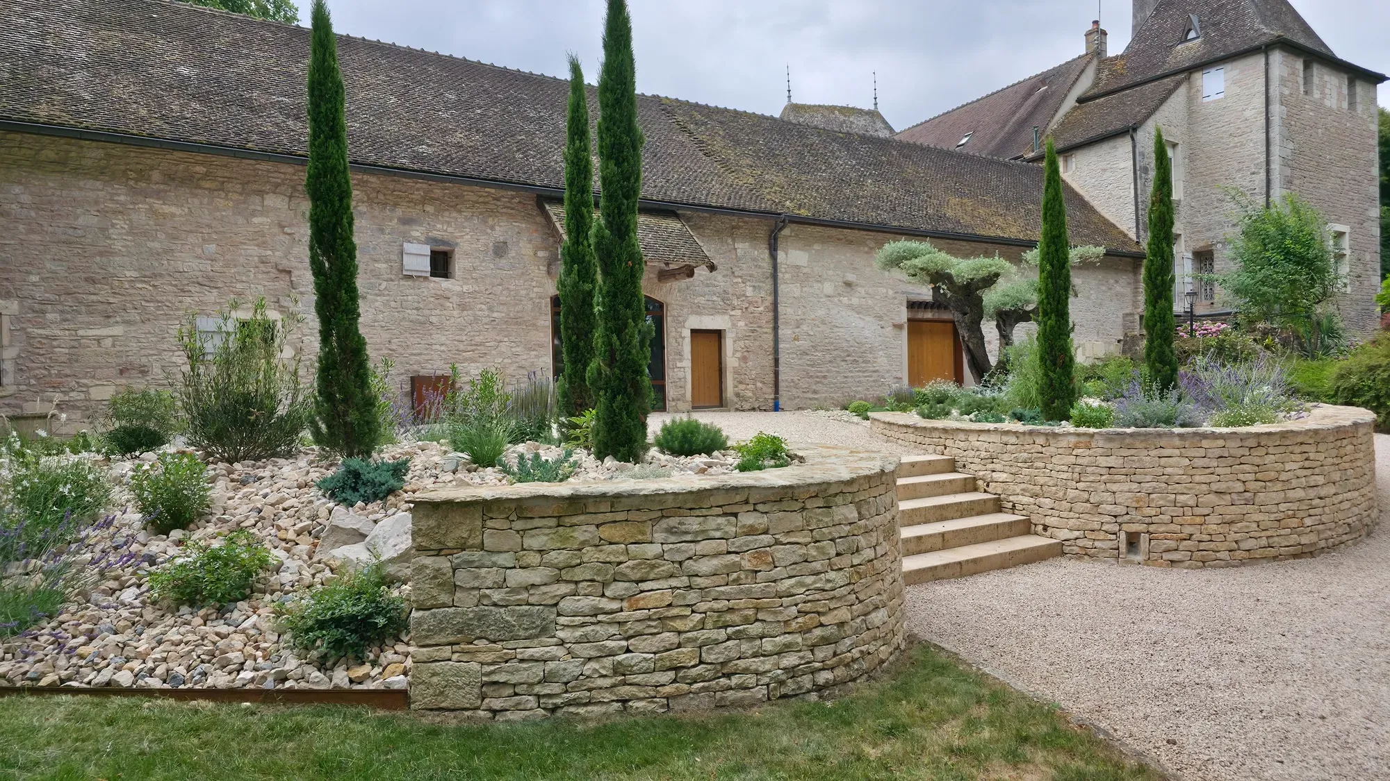 Cour en gravier avec cyprès, massif surélevé en pierre sèche et arbres taillés devant un corps de ferme – Hortiplan Paysagiste