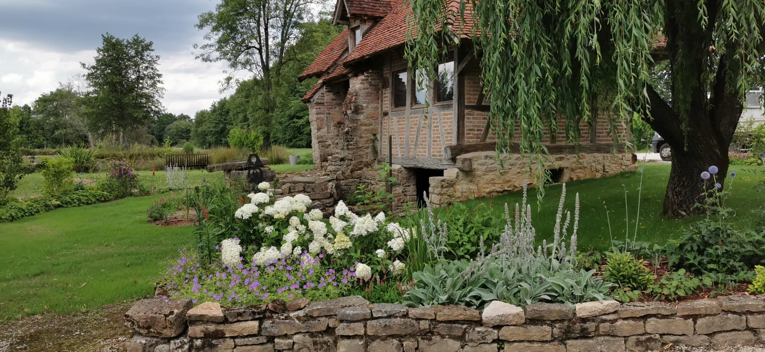 Massifs de vivaces fleuries et muret en pierre devant une maison à colombages – Hortiplan Paysagiste