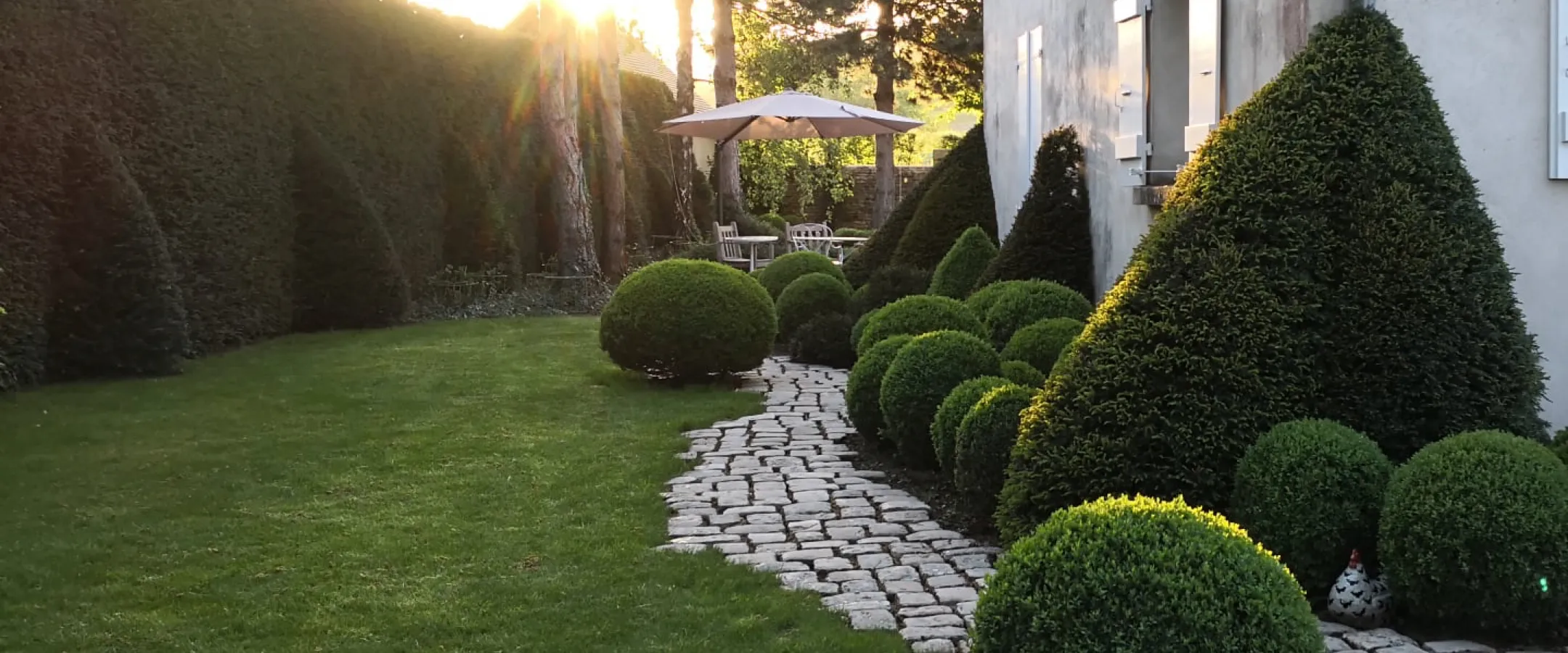 Boules de buis et ifs taillés le long d'une allée pavée sur pelouse au soleil couchant – Hortiplan Paysagiste